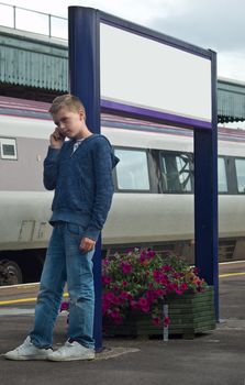 an image of a young teenage boy talkuing on a mobile phone at a train station, leaning against a sign post.