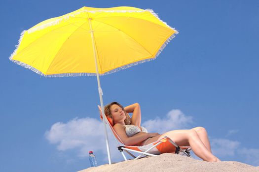 Blond woman in bikini relaxing (sunbathing) under an umbrella on the beach