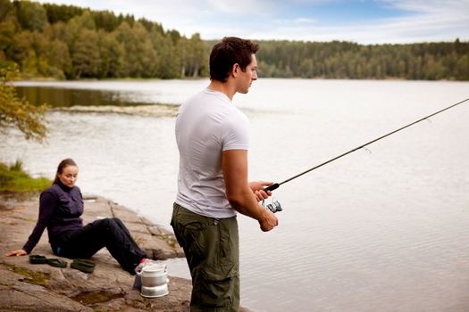 A man fishing on a lake with camping equipment and woman in background