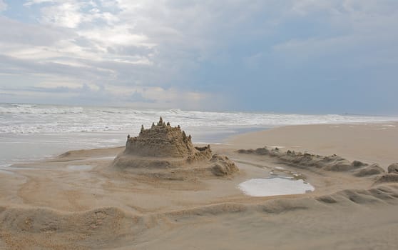 Landscape image of a sand castle with a moat around it.  Ocean and summer sky both included in image.