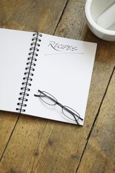 A blank wire spiral bound recipe book with the title 'recipe' hand written at the top of the page. A pair of black framed glasses rest on the page. Set on a wooden kitchen table top with the corner of a white ceramic pestle and mortar visible in the top right hand corner.