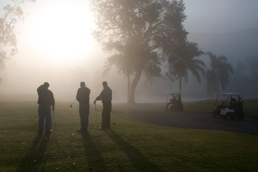 Early morning golfers silhouetted in a dense fog with a rising sun