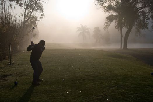 Early morning golfers silhouetted in a dense fog with a rising sun