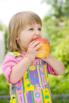 little girl in a multi-coloured dress bites off an apple in the solar afternoon
