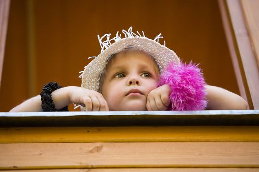 The little girl in a summer hat looks out of a window of a wooden country house
