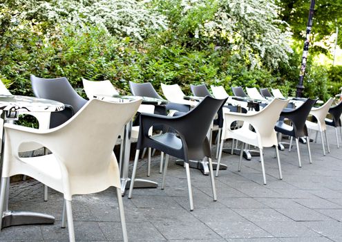 Summer cafe with empty white both black plastic tables and chairs nearby to green bushes
