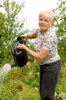 The elderly woman pours water on flowers in the garden in summer day
