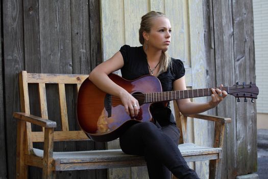 Photo of a beautiful blond female playing her acoustic guitar on an old wooden bench.  