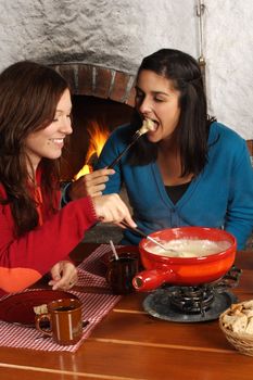 Photo of two beautiful females dipping bread into the melted cheese in a fondue pot. Focus is on girl on the right.