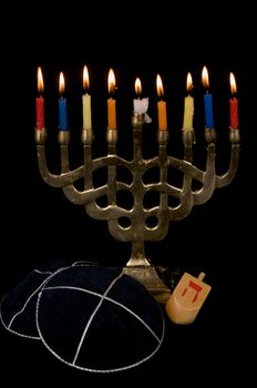 A menorah with 9 burning candles is seen with two yarmalkas and a dreidel, isolated on black