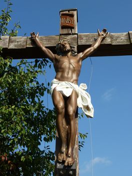 Sculpture of suffering Jesus on a wood cross with green vegetation next to it by beautiful weather