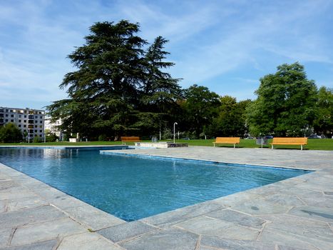 Swimming pool in a park surrounded by tiles, benches and trees by little cloudy weather