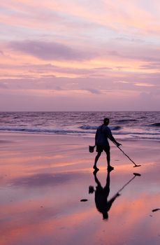 A man using a metal detector on the beach at sunrise.