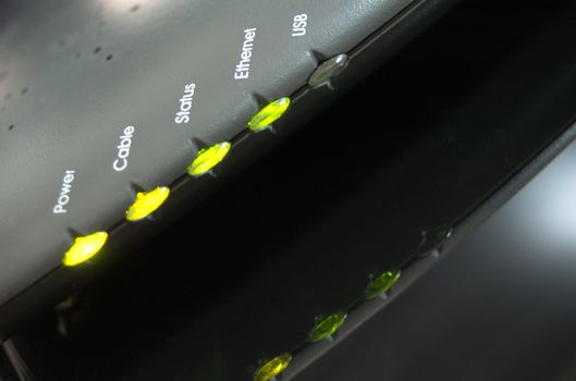 Angled close up on a row of LEDs showing the status of a cable modem. Their reflection can be seen in the black glass table the modem rests on.