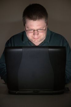 Man works in a darkened room at a laptop. He is illuminated by the laptop screen and is smiling, looking down toward the keyboard.
