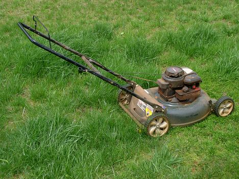 A dusty gasoline-powered push lawnmower stands in long grass which is badly in need of mowing.
