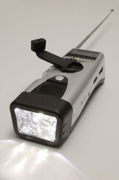 A wind-up weather-band radio with built in flashlight, hand crank and antenna extended, and light switched on, over a mid-gray background.