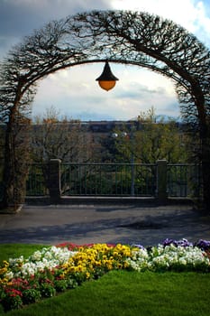 Image of an archway made of vines in a Lausanne park with hanging lamp and flowers.