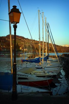 Marina in Lausanne Switzerland showing various boats, sailboats, lamp post and landscaped hills with vineyards in the background.