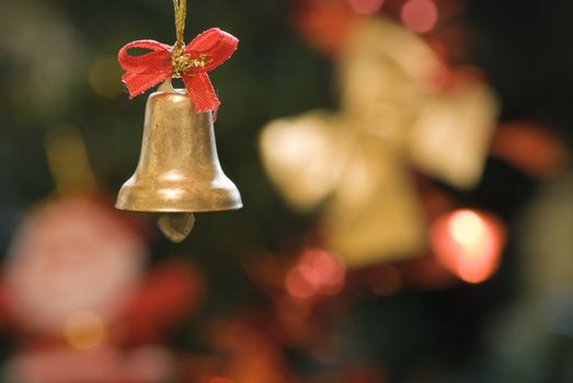 Decorative xmas bell with red ribbon with blurred xmas tree decoration in the background.