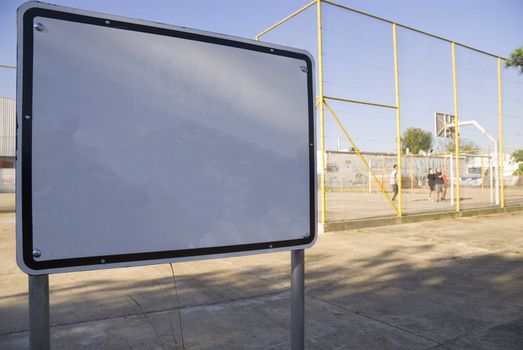 Shot of a blank white metal board with people playing basketball in the far background, in a fenced court. Half-image copy space.