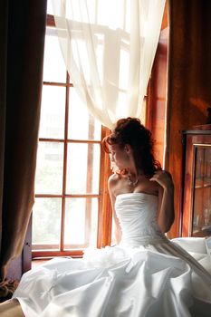 Young adult bride wearing white wedding dress sat by window in sunlight.