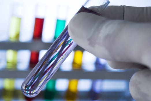 Lab personnel handles test tube, studio shot; line of test tubes containing different colored liquids at background
