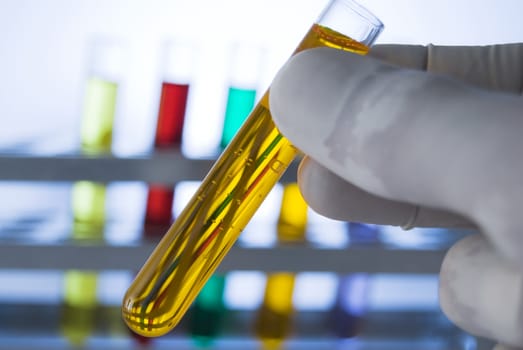 Lab personnel handles test tube, studio shot; line of test tubes containing different colored liquids at background
