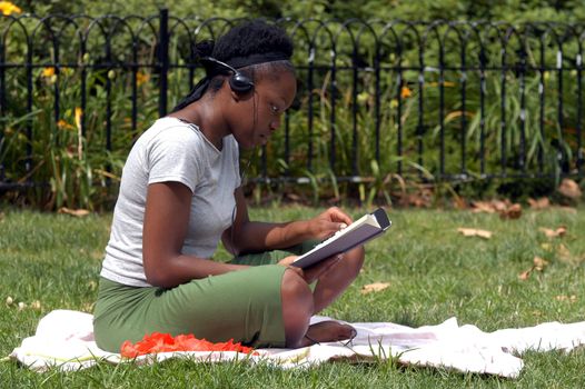 A young african american woman sits in the park and reads while listening to music.