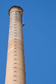 Huge Industrial Chimney with unrecognizable workers hanging from it. Blue sky.