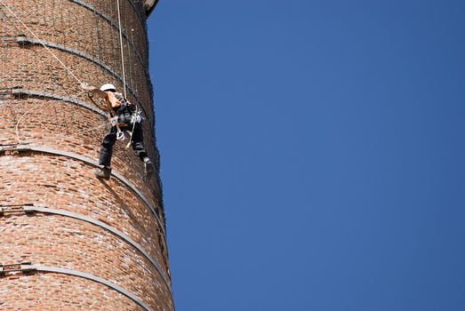 Cropped shot of an unrecognizable workers hanging from a high industrial brick chimney. Blue sky in the background.
