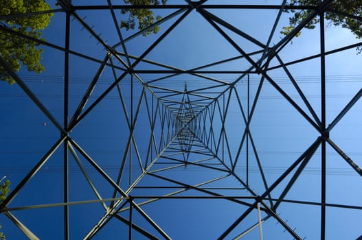 Inside an electricity pylon, surrounded by a few green trees, looking directly up at the clear blue sky. It is as if the pylon forms a metal spider's web.