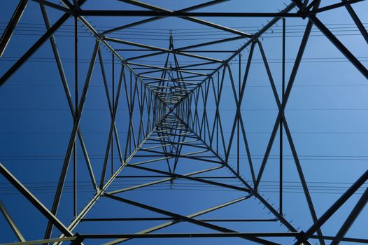Inside an electricity pylon looking directly up at the clear blue sky. It is as if the pylon forms a metal spider's web. A tiny aeroplane seems to be caught in the bottom left corner.