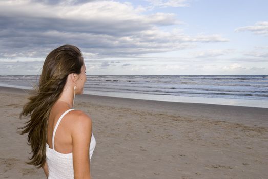 Beautiful young girl in the sand (late afternoon), with cloudy sky, looking out into the sea.