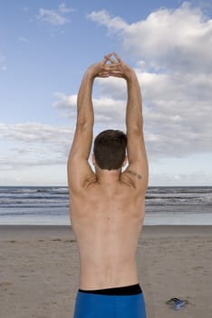 Young man stretching in the beach with cloudy sky. Shot from behind.