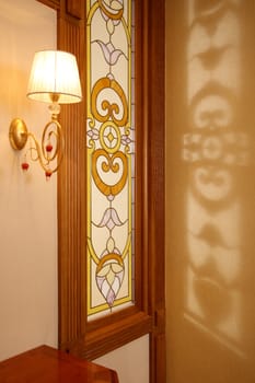 Lamp on a wall with a stained-glass window through which appears through the sun
