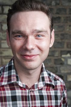 Headshot of young white man wearing checkered shirt and smiling with brick wall in the background.