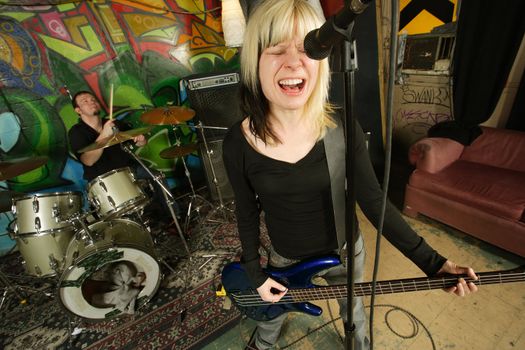 Female bass player screaming into a microphone.  Focus on singers face - drummer blurry in the background.  Shot with slow shutter speed and strobes - motion blur visible in some areas.
