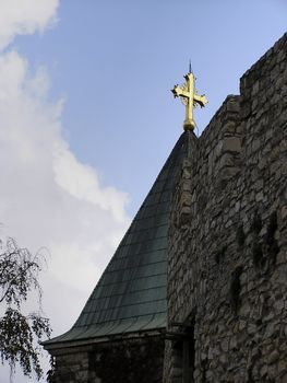 Detail of church roof with golden cross at top near stone wall.