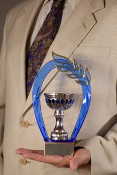 Businessman, dressed in a pale suit, hoding an award in the palm of his hand. Space on the plaque of the award for text.