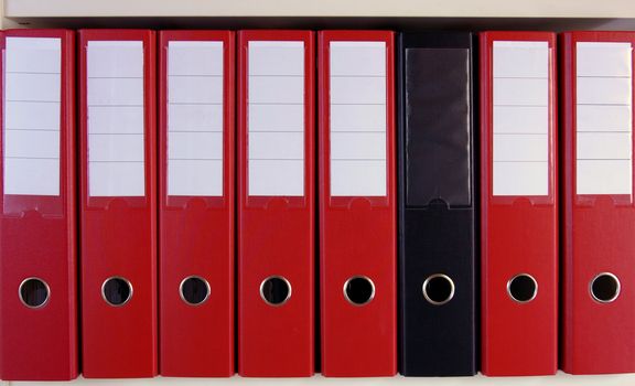 A row of red lever arch files on a shelf, with one black, un-marked one in their midst.