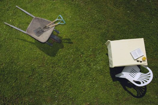 Vertical view of a wheelbarrow, with garden tools, and a garden table with book, sunglasses and drink, all on a green lawn. Space for text on the green of the grass.