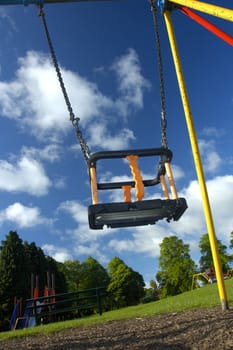An empty swing in a children's playground, taken from a low viewpoint. Other playground equipment, also empty, can be seen in the background.