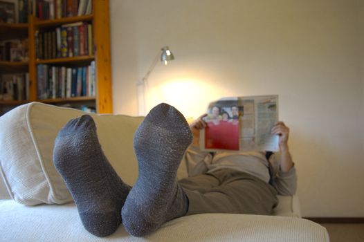 A man lies on the couch with his feet up, reading the paper. Focus on the slightly worn socks. Space for text on the wall, top right.