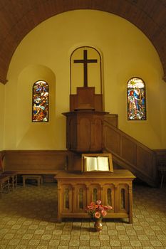 The plain, unadorned, pulpit in a small protestant church in Switzerland. In front of it stands a small wooden altar on which an open bible rests. Lit by the warm glow of a late afternoon sun coming in through the side windows.