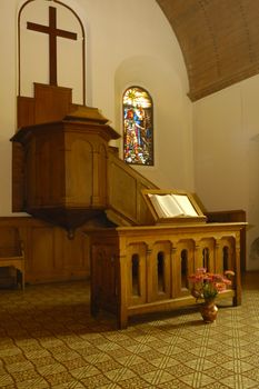 The plain, unadorned, pulpit in a small protestant church in Switzerland. In front of it stands a small wooden altar on which an open bible rests. Lit by the warm glow of a late afternoon sun coming in through the side windows.
