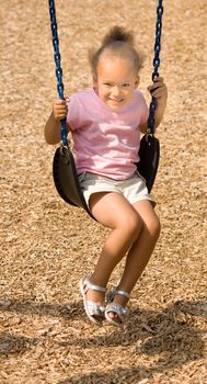 Beautiful little mixed race girl on swing with a smile wearning a pink shirt, shorts and sandals. The scene was set against a playground wood chip mulch textured background.