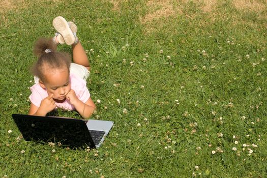 Beautiful young ethnic race girl using laptop on a field of green grass and daisy wheel and clovers. She has the expression of contemplating as she surfs the internet.