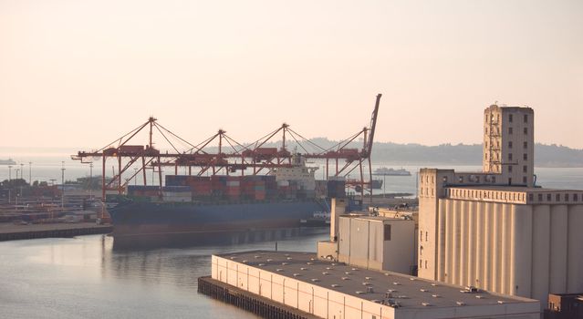 Container Ship docked at the Port of Seattle next to container cranes. Storage Silos are grouped in the foreround. The sun is setting across the waters of the Puget Sound.