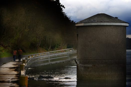 Point Defiance Boardwalk and Landmark is a capture of the long winding boardwalk at Point Defeinace near Tacoma Washington. The large structure in the water has a "high voltage" sign on its door. We think it might be a pump house. 
